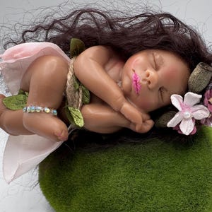 May include: A sleeping baby doll with dark hair, wearing a pink skirt and a beaded ankle bracelet. The doll rests on a green mossy surface, adorned with flowers and leaves. The baby has pink lips and is nestled in a curled position.