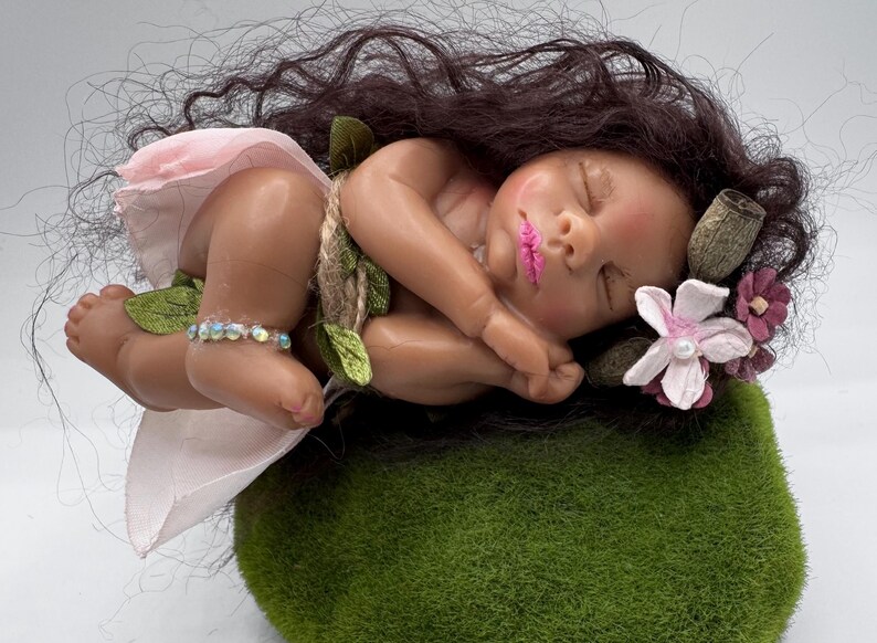 May include: A sleeping baby doll with dark hair, wearing a pink skirt and a beaded ankle bracelet. The doll rests on a green mossy surface, adorned with flowers and leaves. The baby has pink lips and is nestled in a curled position.
