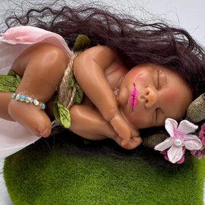 May include: A sleeping baby doll with dark brown hair and pink lips, wearing a pink skirt and a beaded ankle bracelet. The doll is adorned with green leaves and rests on a bed of green moss, with flowers in its hair.