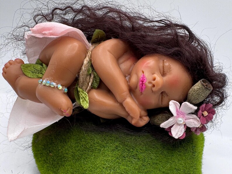 May include: A sleeping baby doll with dark brown hair and pink lips, wearing a pink skirt and a beaded ankle bracelet. The doll is adorned with green leaves and rests on a bed of green moss, with flowers in its hair.