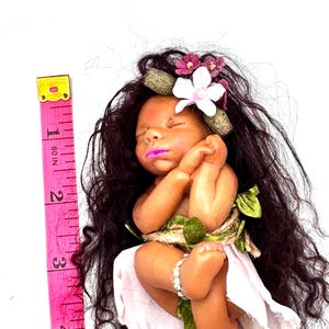 May include: A handcrafted baby doll with dark hair, a floral crown, and a pink dress. The doll is adorned with a bracelet and wrapped with twine and green leaves. A pink measuring tape is placed next to the doll, showing a length of approximately 4 inches.