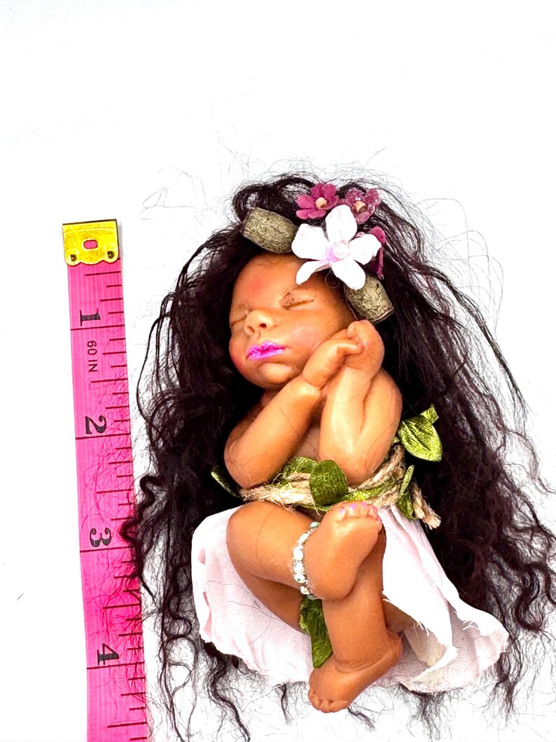 May include: A handcrafted baby doll with dark hair, a floral crown, and a pink dress. The doll is adorned with a bracelet and wrapped with twine and green leaves. A pink measuring tape is placed next to the doll, showing a length of approximately 4 inches.