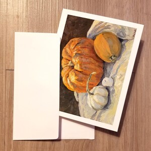 Peut inclure: Une peinture de trois citrouilles sur un tissu blanc. Les citrouilles sont orange et jaunes. La peinture est sur une carte blanche.