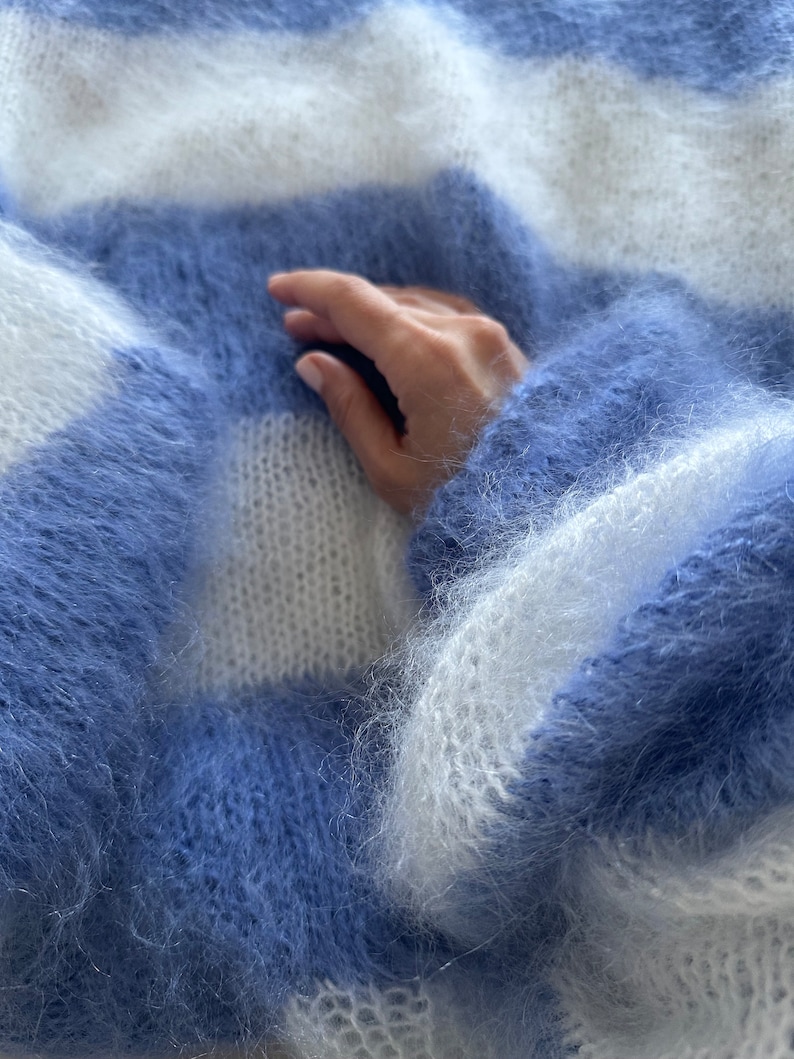 Op de afbeelding: Een zachte, pluche blauwe en witte gestreepte trui met een hand die op de stof rust.