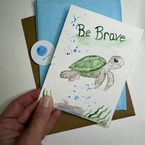 Op de afbeelding: Waterverf wenskaart met een groene zeeschildpad en de tekst "Be Brave" in het groen. De kaart wordt in een hand gehouden, met blauw vloeipapier en een bruine envelop op de achtergrond. Een kleine ronde sticker is ook zichtbaar.