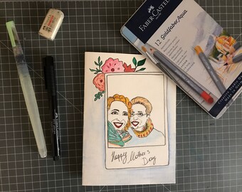 May include: A handmade card with a watercolor illustration of two women smiling. The card has a floral border and the text "Happy Mother's Day".