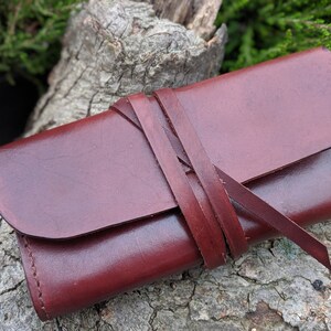 May include: A burgundy leather wallet with a strap closure. The wallet is closed and lying on a piece of tree bark.