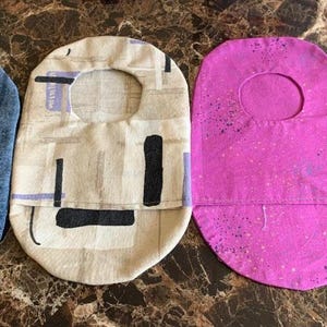 May include: Four fabric bibs with different patterns. One is blue denim, one is beige with black geometric shapes, one is pink with white speckles, and one is purple with red circles.