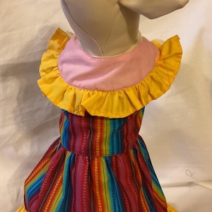 May include: A white stuffed dog wearing a colorful striped dress with yellow ruffles. The dress has a pink collar and a yellow ruffle trim.