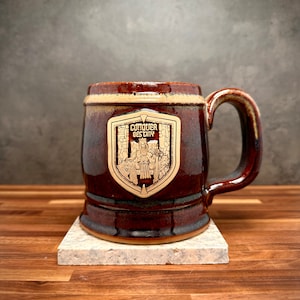 May include: A dark brown ceramic mug with a glossy finish and a sturdy handle. Centered on the front is a light-colored shield-shaped emblem featuring an illustration of a seated figure with a sword and the text "CONQUER DESTINY" in bold letters.