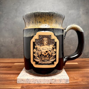 May include: A black ceramic mug with a tan rim and a tan rectangular emblem on the front. The emblem features a graphic of a bottle, a skull, and floral elements with the text "The Dose Makes The POISON" printed in black lettering.