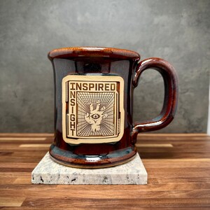 May include: A glossy dark brown ceramic mug with a handle and a light-colored rectangular patch on the front. The patch features a black line drawing of a hand with an eye in the palm, set against a grid background, with the text INSPIRED INSIGHT.