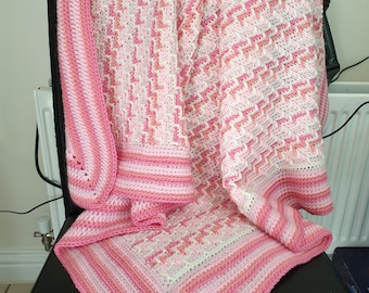 May include: A pink and white crocheted baby blanket with a striped border. The blanket is folded and draped over a black chair.