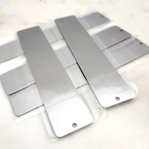 May include: Four silver metal bookmarks with rounded corners and a hole at the top. The bookmarks are packaged in clear plastic.
