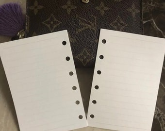 May include: Two white lined paper refills for a personal planner with six holes on each side. The refills are laying on a brown leather planner with a monogram pattern.