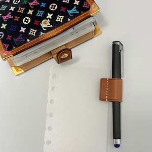 May include: A white planner page with a brown leather pen holder and a black pen with a purple tip. The planner page has holes on the left side for a binder.