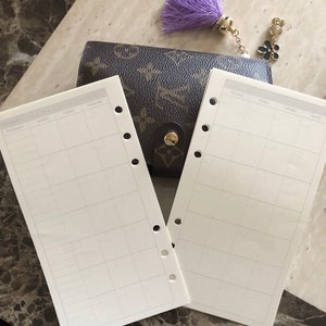 May include: A brown patterned wallet with a gold button and a purple tassel charm. Two blank white planner pages with gray grid lines are in front of the wallet, ready for use.