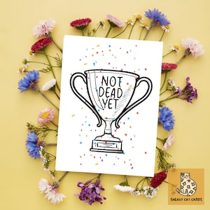 May include: A white greeting card with a black and white trophy illustration and the text "NOT DEAD YET". The card is surrounded by colorful flowers and confetti on a yellow background. The card is from Sneaky Cat Cards.