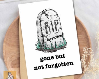 May include: A white greeting card with a tombstone illustration that reads "RIP twenties" and the text "gone but not forgotten". The card is on a wooden tray with a brown envelope.