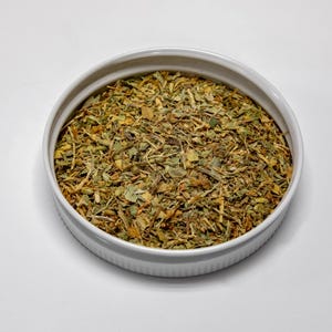 May include: A white ceramic bowl filled with a mix of dried herbs and spices. The mixture includes green, brown, and yellow elements, suggesting a blend for culinary use. The bowl is round and sits on a white surface.
