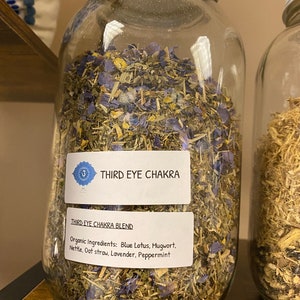 Op de afbeelding: Een glazen pot gevuld met een mengsel van gedroogde kruiden voor de derde oog chakra. Het etiket vermeldt "Third Eye Chakra Blend" en somt de biologische ingrediënten op: Blauwe Lotus, Bijvoet, Brandnetel, Haverstro, Lavendel, Pepermunt.