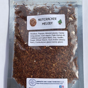 Puede incluir: Una bolsa de plástico transparente llena de una mezcla de té de hojas sueltas. El té es una mezcla de trozos marrones y blancos. La bolsa tiene una etiqueta que dice "NUTCRACKER MELODY" y enumera los ingredientes: Rooibos, papaya, trozos de almendras, trozos de caramelo, fécula de maíz, aceite de palmiste, goma de celulosa (fibra vegetal), soja, vainillina, azúcar, fécula de trigo, goma arábiga (fibra dietética), glaseado de confitería (glaseado de caramelo). La etiqueta también incluye el nombre de la empresa y el sitio web: AWAKEN ONE HOME REMEDIES LLC, https://www.awakenonehomeremedy.com/.