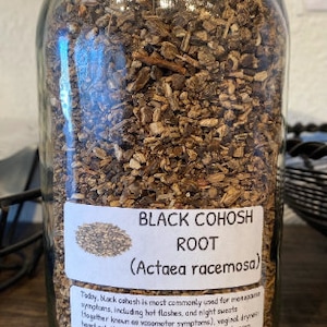 Op de afbeelding: Een glazen pot gevuld met gedroogde zwarte cohosh-wortel. Het etiket vermeldt "Black Cohosh Root (Actaea racemosa)" en somt de gebruikelijke toepassingen van het kruid op, waaronder opvliegers en nachtelijk zweten.