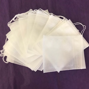 Könnte beinhalten: Ein Stapel weißer, quadratischer Teebeutel mit angebrachten Schnüren, angeordnet auf einer dunkelvioletten Oberfläche. Die Teebeutel bestehen aus feinem, porösem Material.