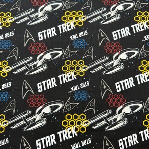 K&ouml;nnte beinhalten: Schwarzer Stoff mit einem sich wiederholenden Muster aus dem Star Trek-Logo, dem Raumschiff Enterprise und Wabenformen in Gelb, Rot und Blau.