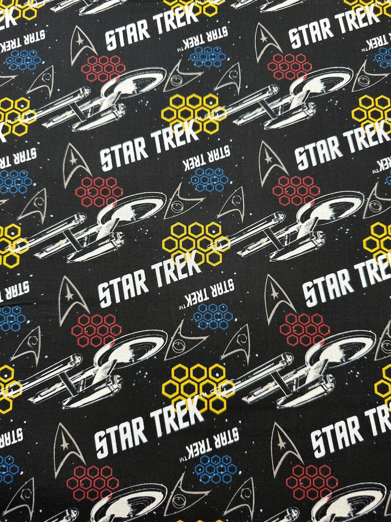 K&ouml;nnte beinhalten: Schwarzer Stoff mit einem sich wiederholenden Muster aus dem Star Trek-Logo, dem Raumschiff Enterprise und Wabenformen in Gelb, Rot und Blau.