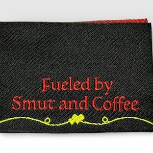 May include: Black fabric label with red embroidered text reading "Fueled by Smut and Coffee." A yellow and green embroidered design is at the bottom. The label has a red border.