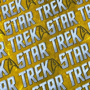 May include: A yellow fabric with a repeating pattern of the words "Star Trek" and the Star Trek logo in silver.