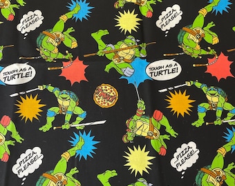 May include: Black fabric with a repeating pattern of Teenage Mutant Ninja Turtles characters, pizza slices, and speech bubbles with the text "...PIZZA, PLEASE!" and "TOUGH AS A TURTLE!"