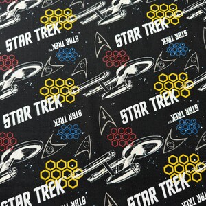 K&ouml;nnte beinhalten: Schwarzer Stoff mit einem sich wiederholenden Muster aus dem Star Trek-Logo, einem Raumschiff und einem Wabenmuster. Der Text "STAR TREK" erscheint mehrmals.