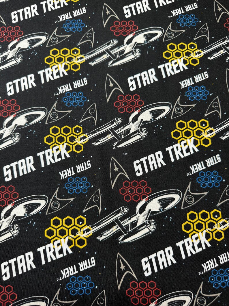 K&ouml;nnte beinhalten: Schwarzer Stoff mit einem sich wiederholenden Muster aus dem Star Trek-Logo, einem Raumschiff und einem Wabenmuster. Der Text "STAR TREK" erscheint mehrmals.