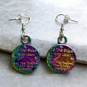 May include: A pair of iridescent metal earrings. Each earring features a circular pendant with the text "To The Stars Who Listen And The Dreams That Are Answered" and a mountain and star design.