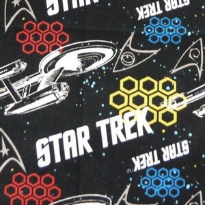 K&ouml;nnte beinhalten: Schwarzer Stoff mit einem sich wiederholenden Muster aus wei&szlig;en Raumschiffen, wei&szlig;em Text mit der Aufschrift "STAR TREK" und gelben, roten und blauen Wabenformen.