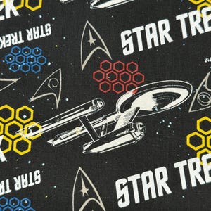 K&ouml;nnte beinhalten: Schwarzer Stoff mit einem sich wiederholenden Muster aus dem Star Trek-Logo, Raumschiffen und dem Starfleet-Abzeichen. Der Hintergrund ist schwarz mit wei&szlig;en Sternen. Das Muster ist in den Farben Gelb, Rot und Blau gehalten.