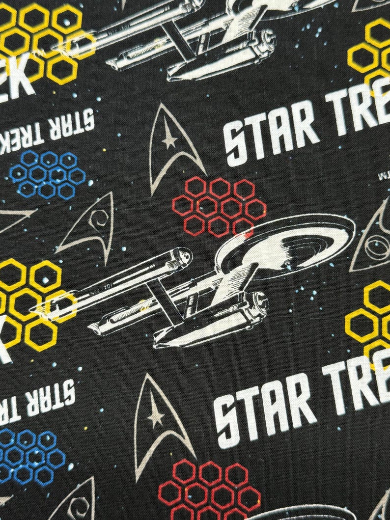 K&ouml;nnte beinhalten: Schwarzer Stoff mit einem sich wiederholenden Muster aus dem Star Trek-Logo, Raumschiffen und dem Starfleet-Abzeichen. Der Hintergrund ist schwarz mit wei&szlig;en Sternen. Das Muster ist in den Farben Gelb, Rot und Blau gehalten.