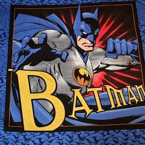 May include: A square fabric panel featuring a graphic illustration of Batman in a grey suit with a blue cape and cowl. The character is posed with a reaching hand and a clenched fist. The word BATMAN is printed in large yellow letters.