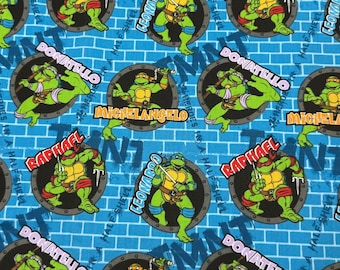May include: Blue fabric with a repeating pattern of Teenage Mutant Ninja Turtles characters. Each turtle is in a circle with their name written below. The names are Leonardo, Donatello, Raphael, and Michelangelo. The background is a blue brick pattern with the word "TMNT" written in white.