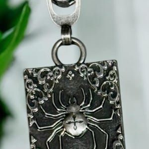 May include: A silver-toned zip pull with a rectangular pendant featuring a spider design. The pendant has an ornate border and a detailed spider illustration. The zip pull is attached to a metal zip.