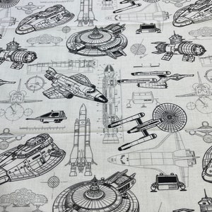 May include: A white fabric with black line drawings of various spacecraft, including rockets, shuttles, and flying saucers. The fabric is a repeating pattern of these spacecraft.