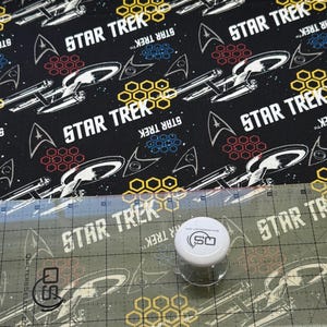 K&ouml;nnte beinhalten: Schwarzer Stoff mit einem sich wiederholenden Muster aus dem Star Trek-Logo, Sternen und Raumschiffen. Der Stoff wird mit einem Lineal und einem Quilting-Werkzeug gemessen.