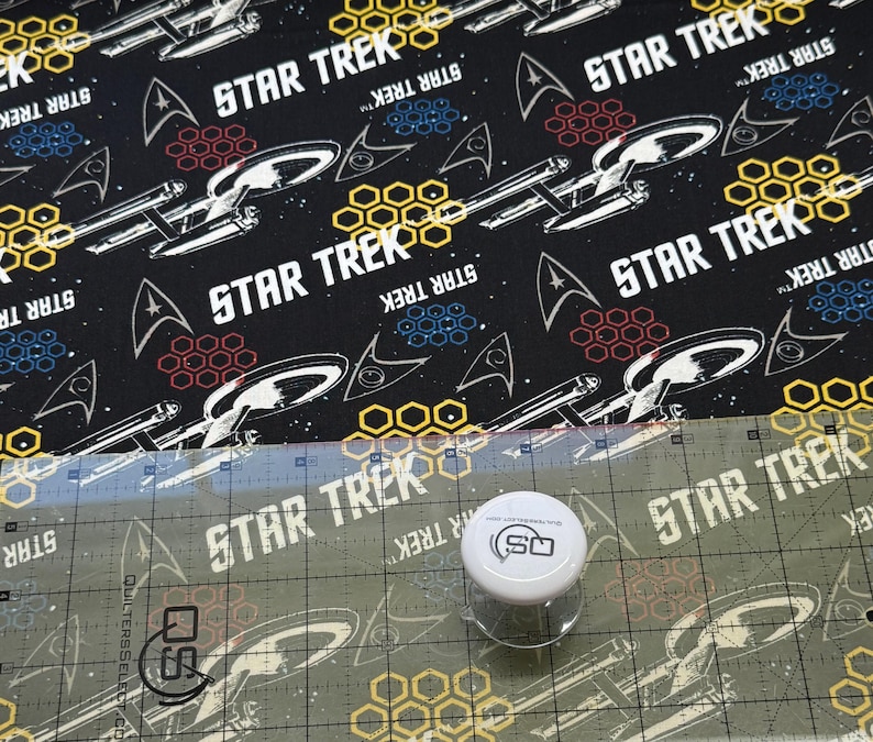 K&ouml;nnte beinhalten: Schwarzer Stoff mit einem sich wiederholenden Muster aus dem Star Trek-Logo, Sternen und Raumschiffen. Der Stoff wird mit einem Lineal und einem Quilting-Werkzeug gemessen.