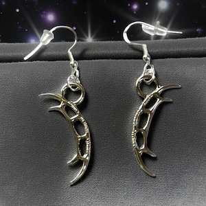 May include: A pair of silver crescent moon earrings. Each earring has a curved, openwork design with three points and a hook for pierced ears. The earrings are displayed on a dark grey surface.