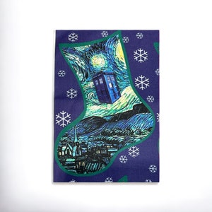 Peut inclure: Une chaussette de Noël bleue et verte avec un motif de nuit étoilée et un TARDIS bleu volant dans le ciel. La chaussette est décorée de flocons de neige blancs.