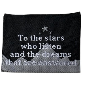 Puede incluir: Un parche rectangular negro con texto bordado blanco y detalles de estrellas. El texto dice "To the stars who listen and the dreams that are answered". La parte inferior presenta un diseño de cordillera.