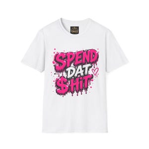 May include: A white short-sleeve t-shirt featuring a graphic print on the chest with the text SPEND DAT $HIT in a stylized, dripping pink and white font. The design includes pink paint splatters and a small pink heart outline on the right side.