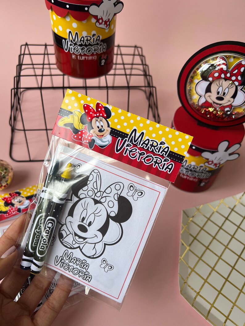 May include: A party favor bag with a Minnie Mouse coloring page and two crayons. The bag has a red, yellow, and white Minnie Mouse design with the text "Maria Victoria" on it.